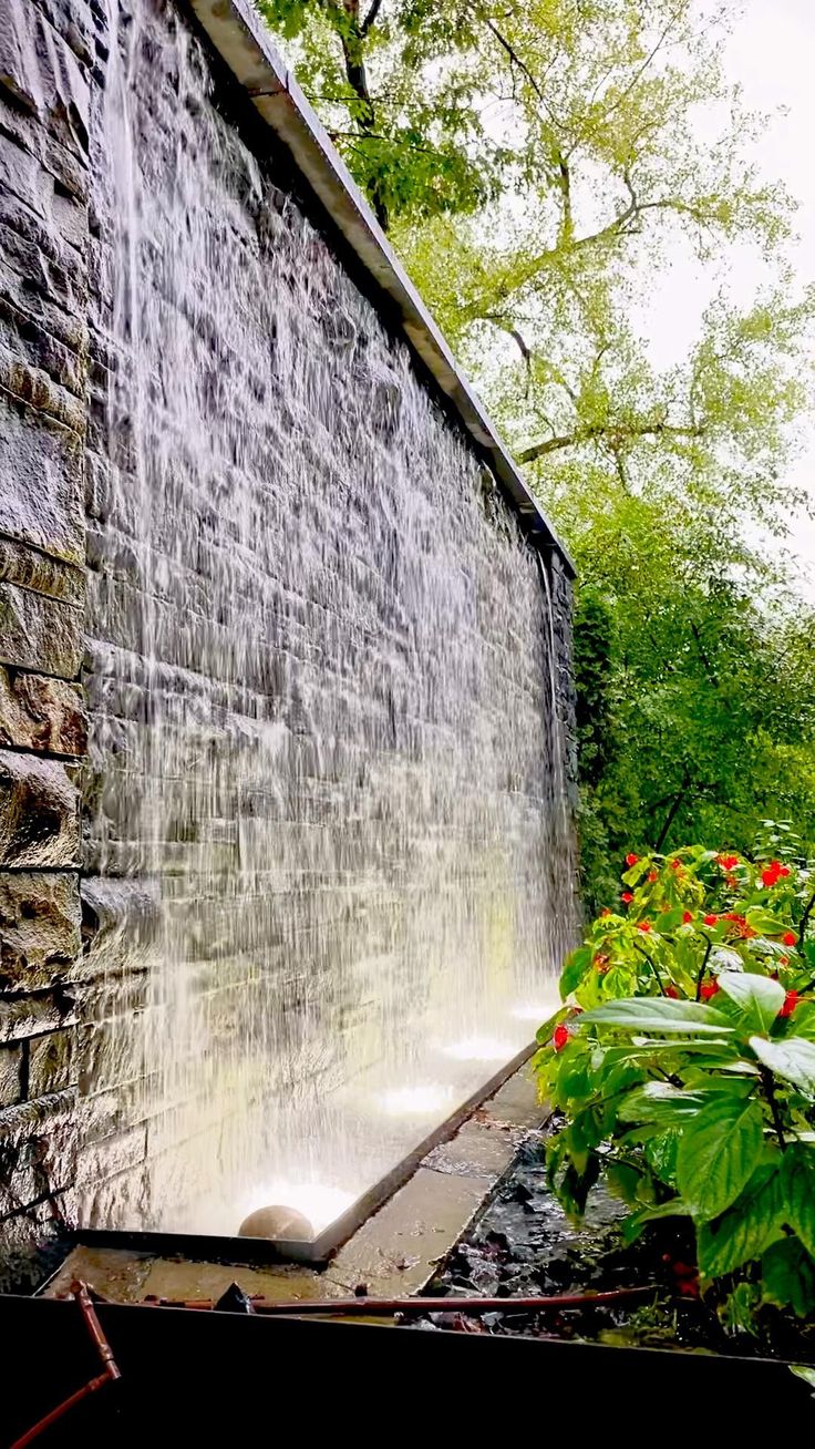 an old brick building with water coming out of it's side and flowers growing in the foreground