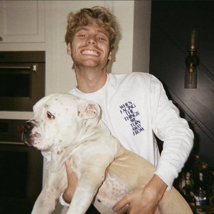 a man holding a white dog in his arms and smiling at the camera while standing next to an oven