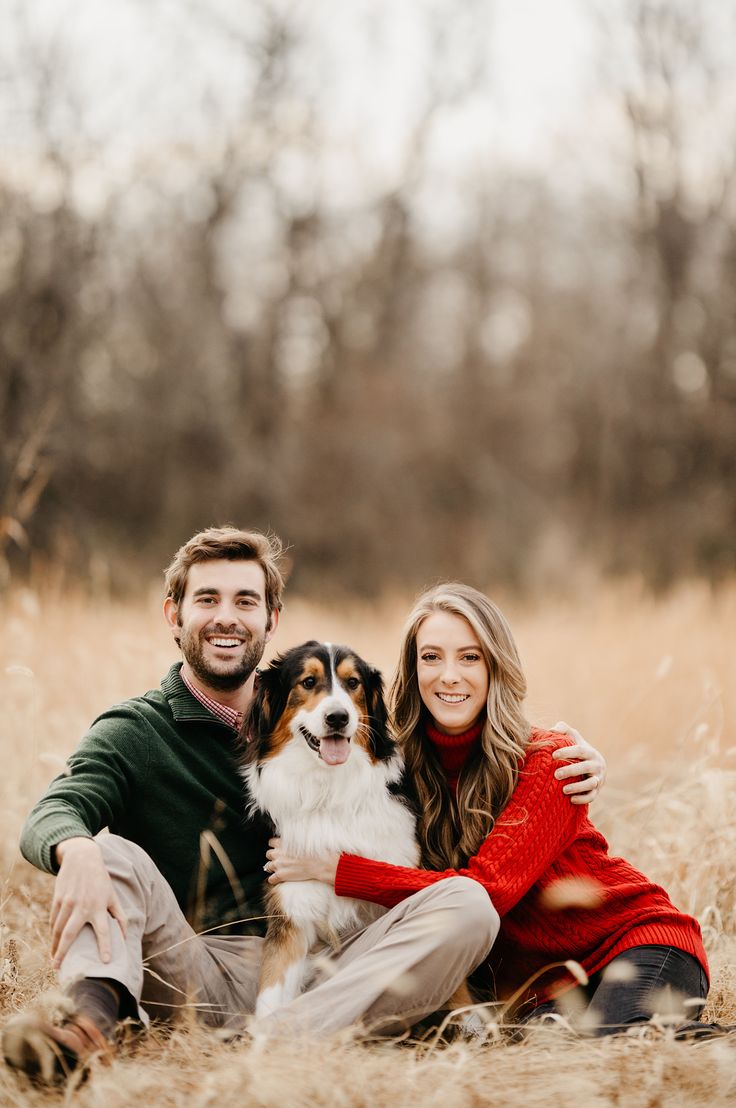 a man and woman are sitting in the grass with their dog, who is wearing a red sweater