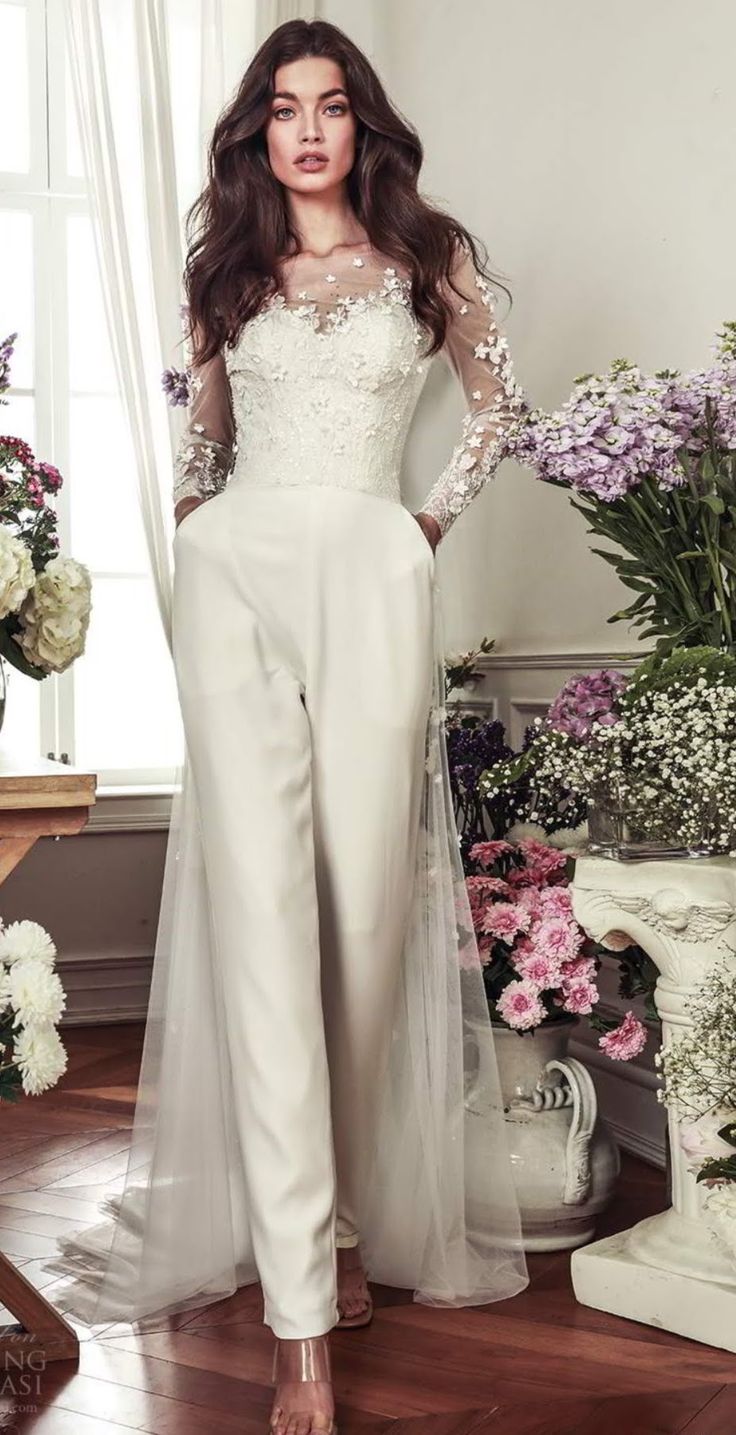 a woman standing in front of flowers wearing a white jumpsuit with sheer sleeves and lace