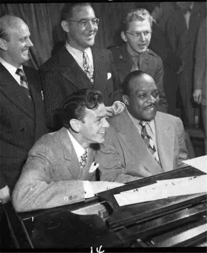 a group of men standing around a piano in front of a man wearing a suit and tie