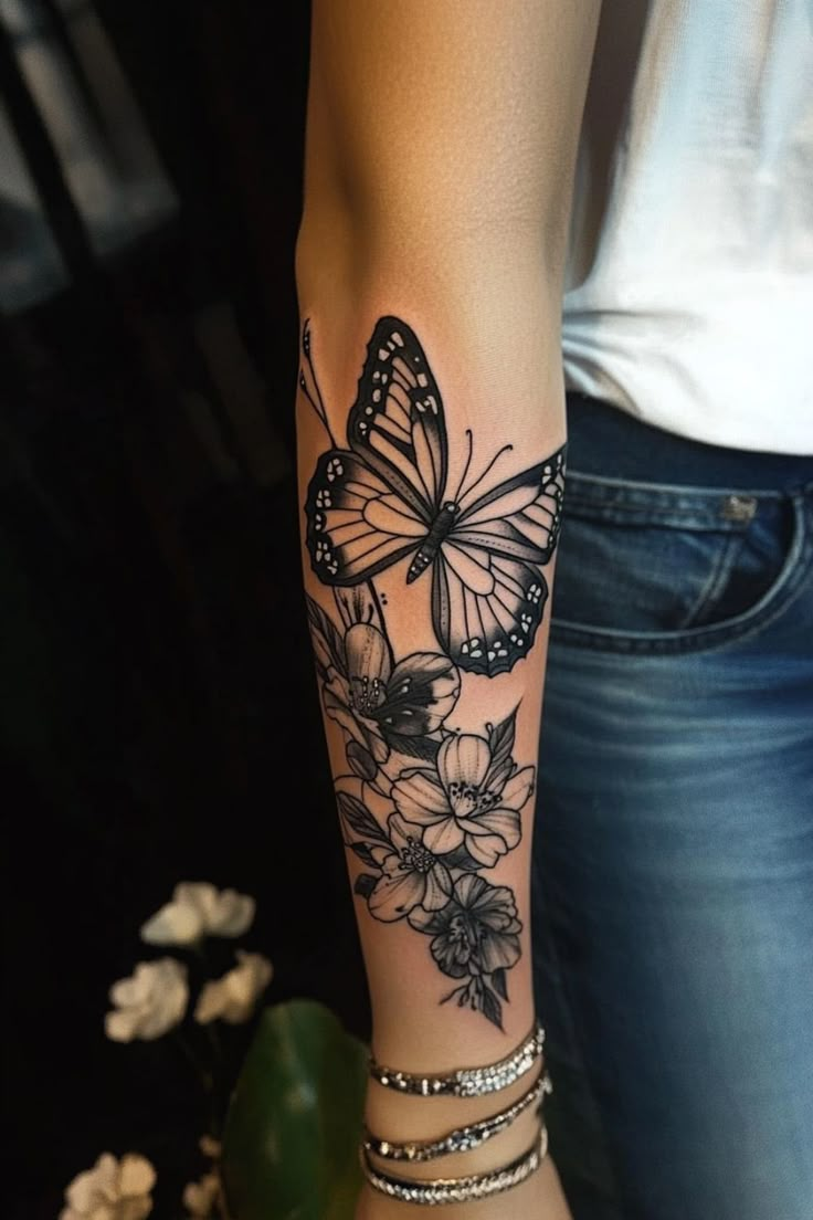 Forearm tattoo of a butterfly and flowers in black ink. Cover Up A Butterfly Tattoo, Shoulder Butterfly Tattoos For Women, Floral And Butterfly Sleeve Tattoo, Butterfly And Flower Tattoo Sleeve, Butterfly Tattoo Ideas For Women, Flower And Butterfly Tattoo, Butterfly Tattoo Designs For Women, Butterfly Flower Tattoo, Skull Butterfly Tattoo