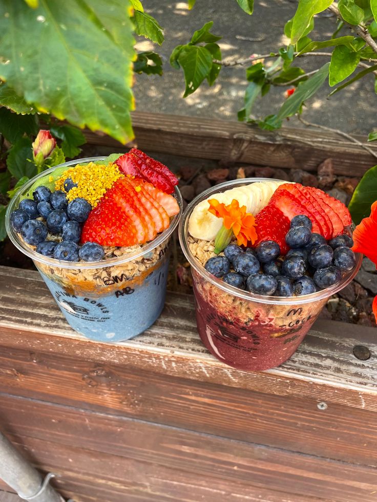 two plastic cups filled with different types of fruit on top of a wooden table next to flowers