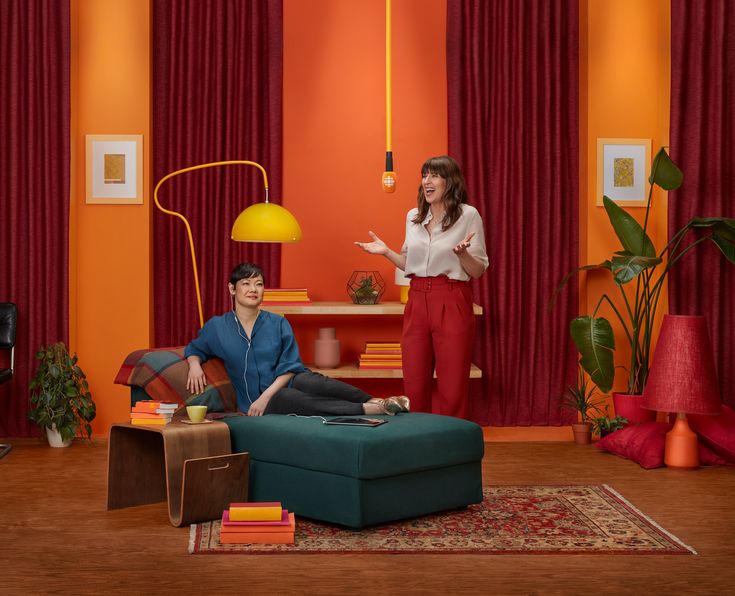 two women are talking in the living room with orange walls and red drapes on the windows