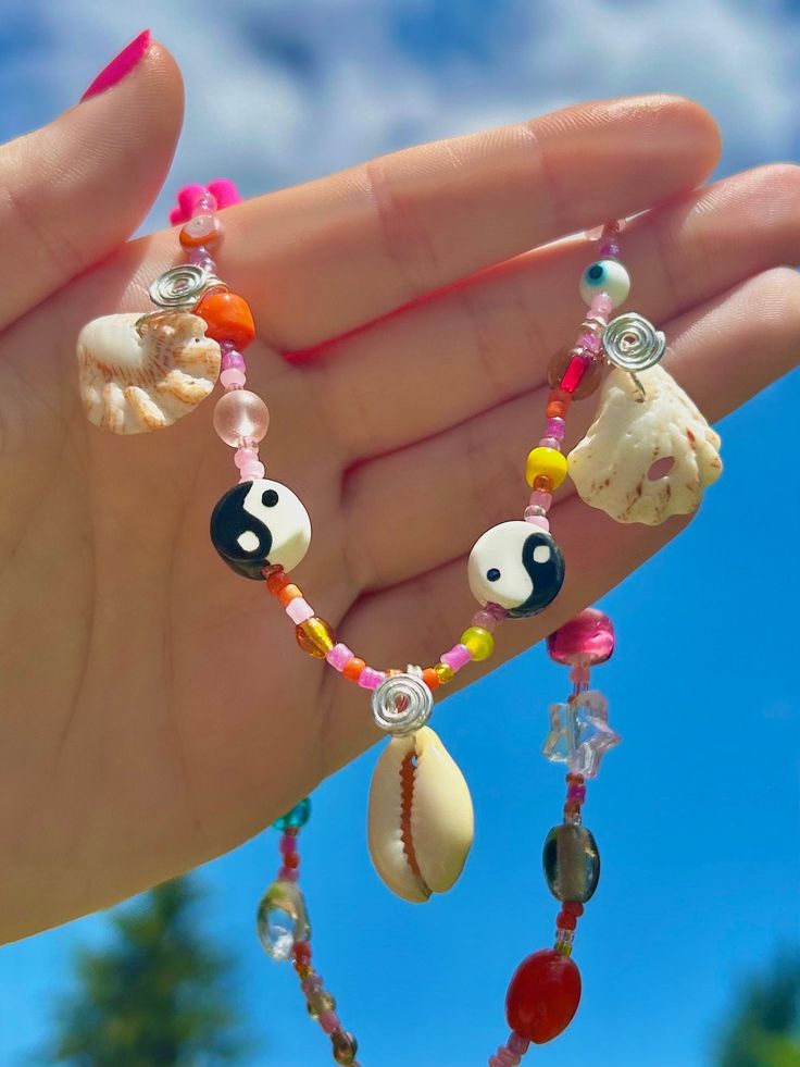 a hand holding a beaded necklace with seashells and charms on it's fingers