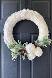 a white wreath hanging on the front door with pine cones and evergreen leaves around it