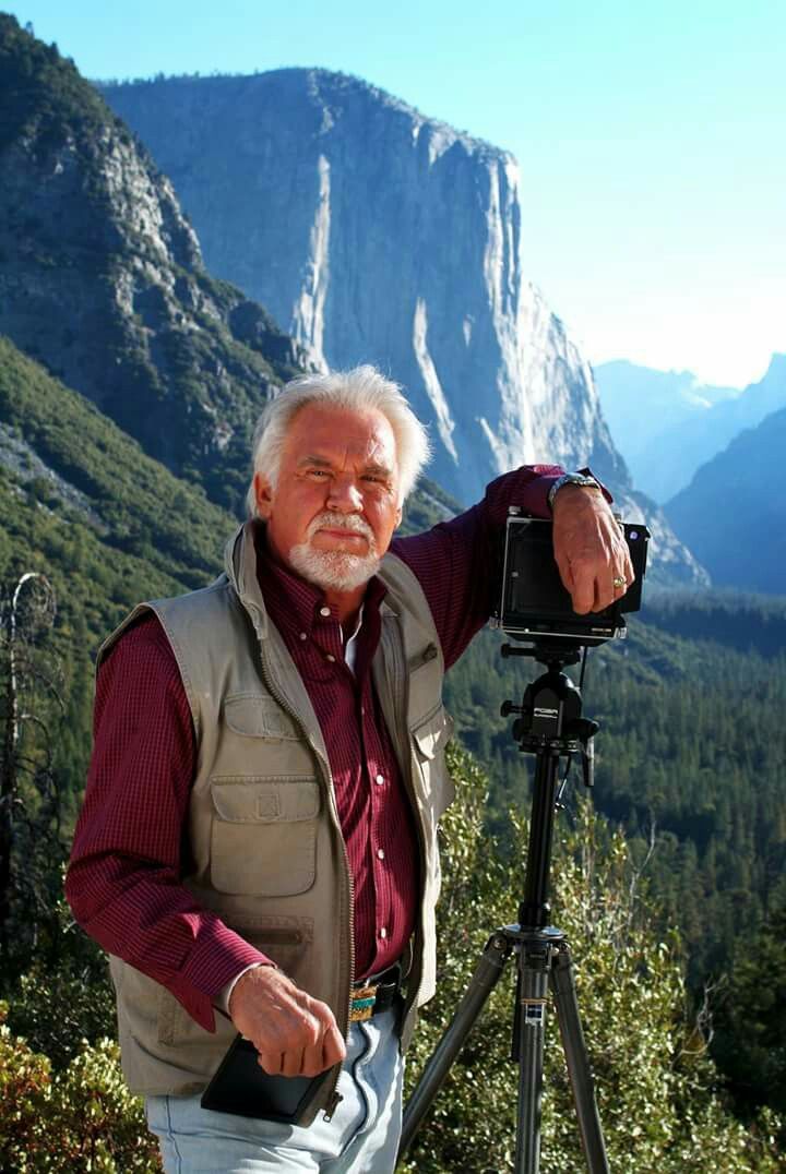 an older man standing in front of a camera on top of a mountain