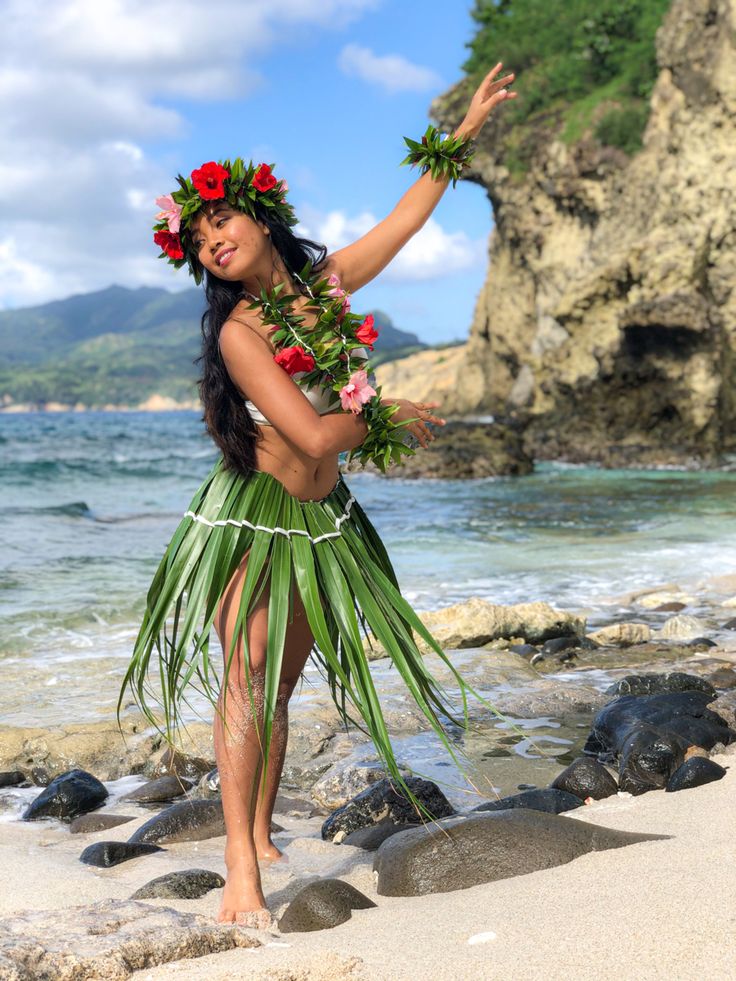 a beautiful young woman standing on top of a beach next to the ocean wearing a hula skirt