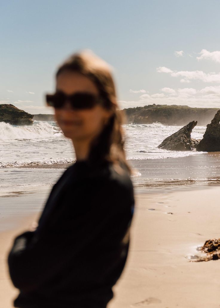 a woman standing on top of a sandy beach next to the ocean with rocks in the background