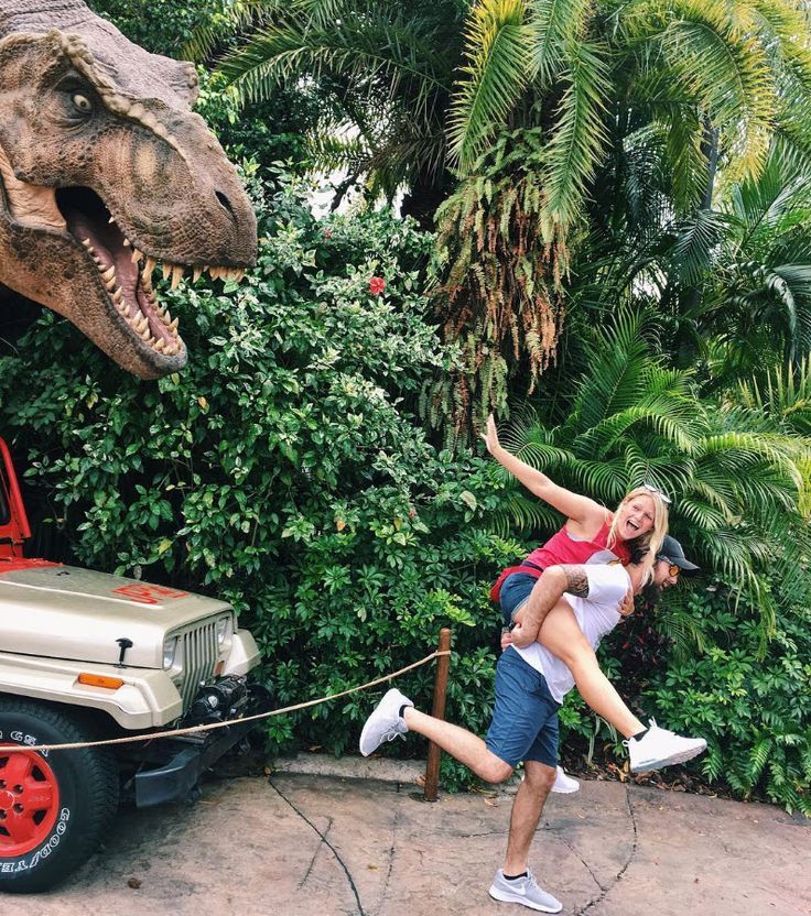 a man and woman are posing in front of a dinosaur statue with a jeep behind them