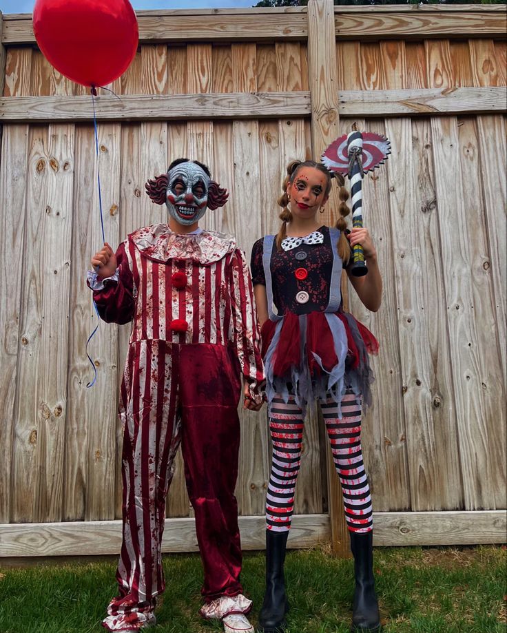 two people dressed as clowns holding balloons in front of a wooden fence with a red balloon