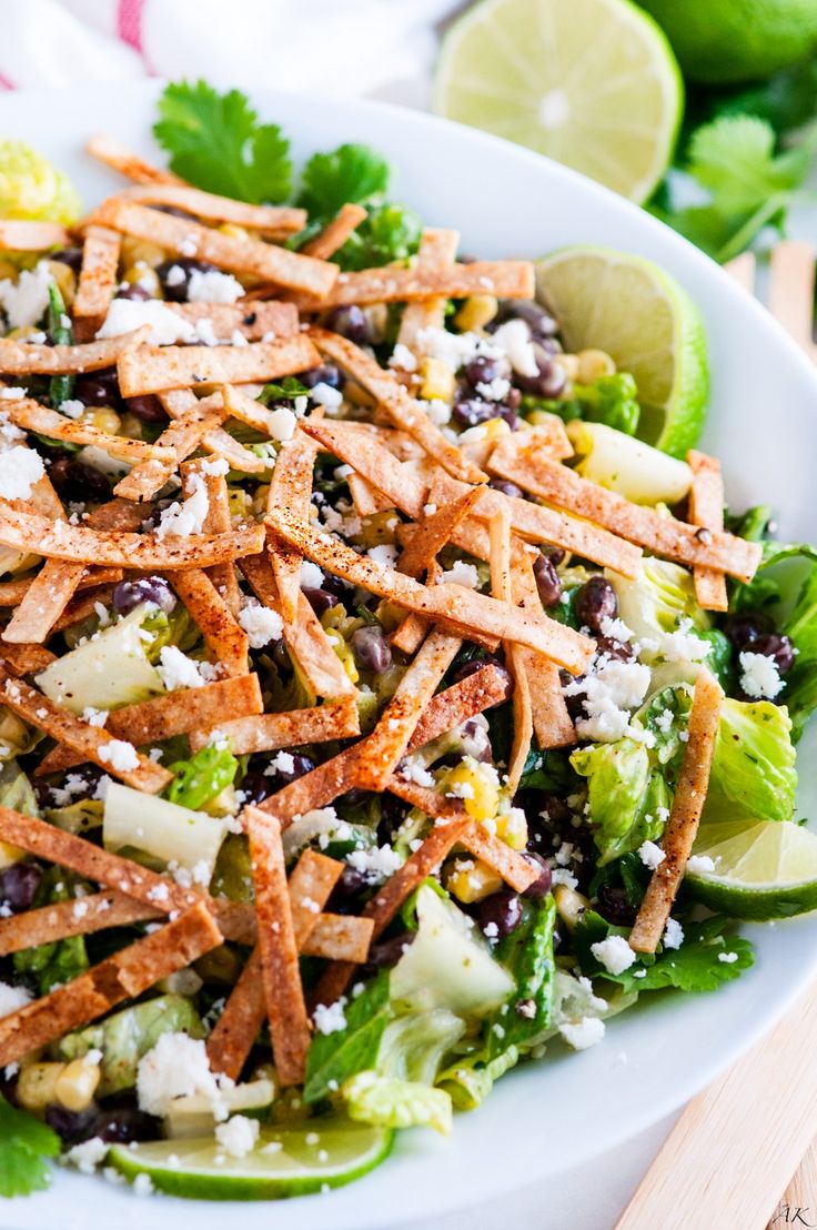 a white bowl filled with salad and topped with chicken, lettuce, black beans, cheese and lime wedges