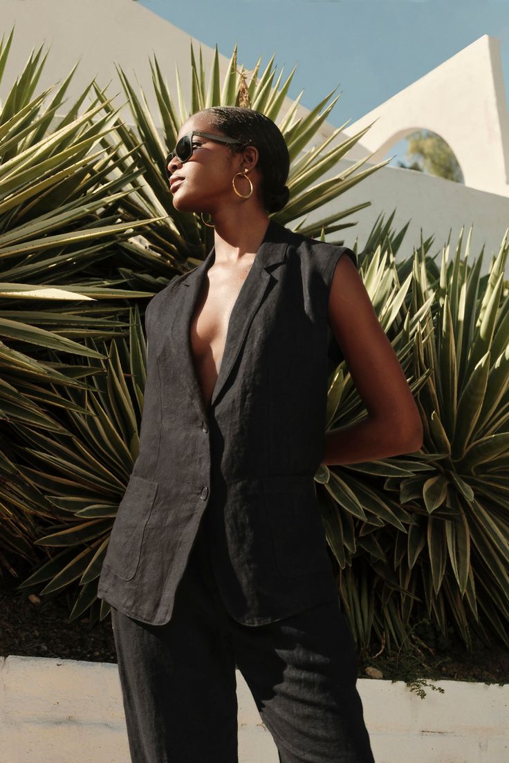 A stylish woman in a black BETHAN HEAVY LINEN SLEEVELESS BLAZER by Velvet by Graham & Spencer stands confidently against a background of sharp green leaves Blazer Linen, Velvet Tees, Sleeveless Blazer, Linen Blazer, Black Blazers, Patch Pocket, Jackets & Coats, Velvet, Blazer