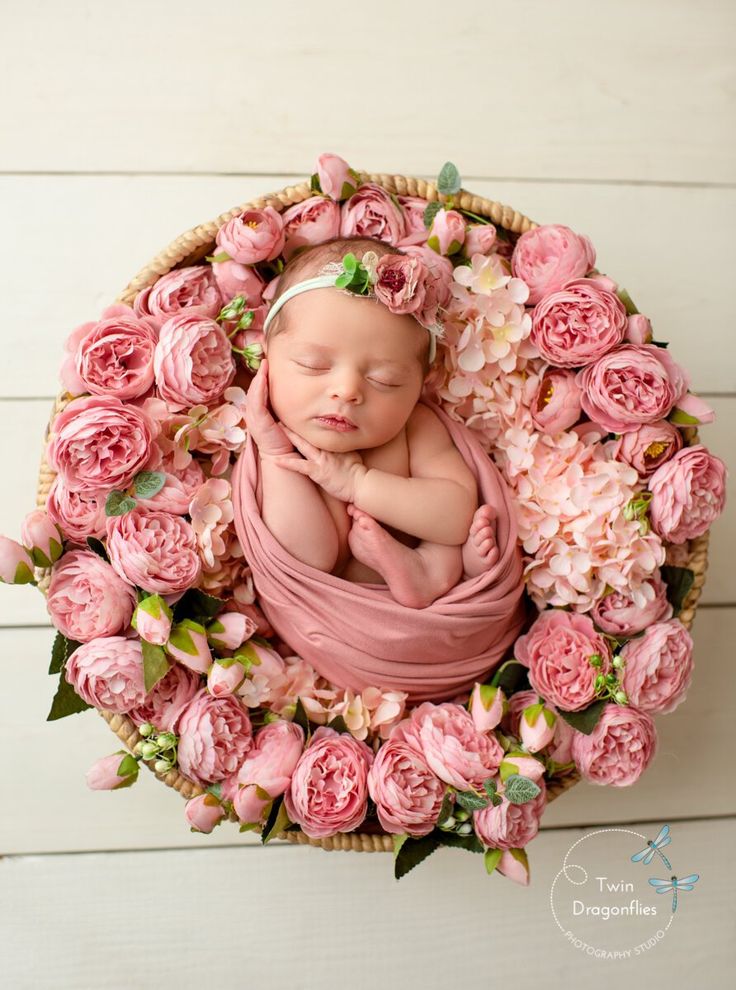 a newborn baby wrapped in a blanket surrounded by pink flowers and greenery on a white wooden background