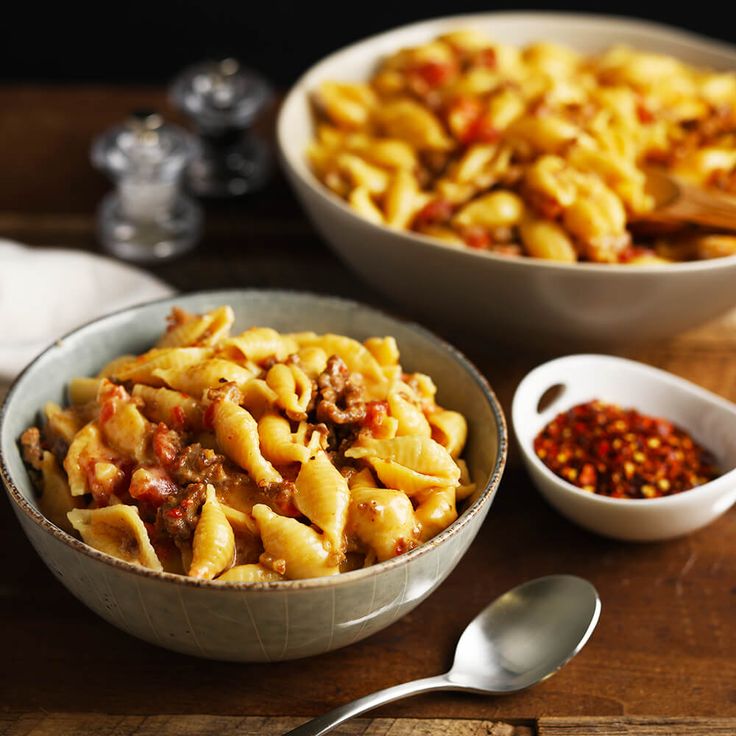 two bowls filled with pasta and meat next to some seasonings on a table top