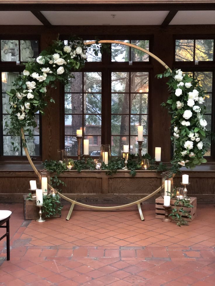 a table with candles and flowers on it in front of large windows, surrounded by greenery