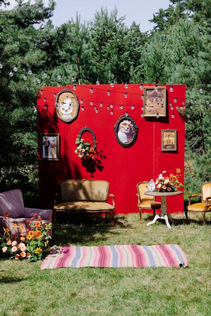 a red wall with pictures on it in the grass next to some chairs and tables