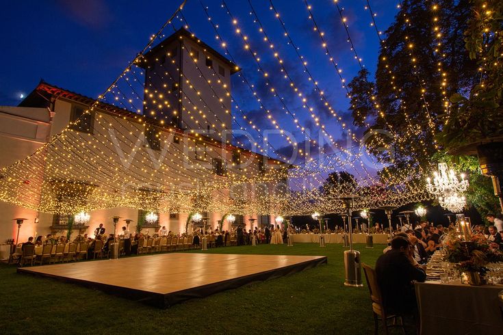 people are sitting at tables in an outdoor area with lights strung from the ceiling and on the ground