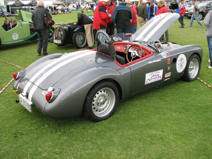 an old race car is parked on the grass with people looking at cars in the background