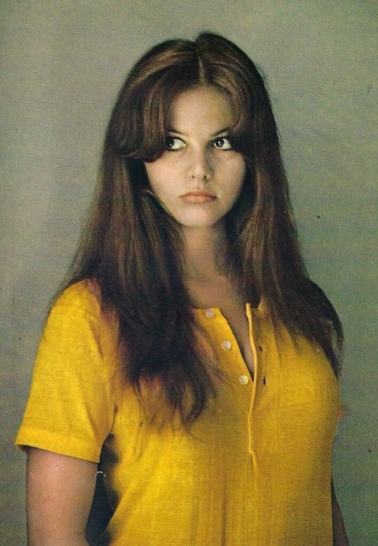 a woman with long brown hair wearing a yellow shirt and looking at the camera while standing in front of a white wall