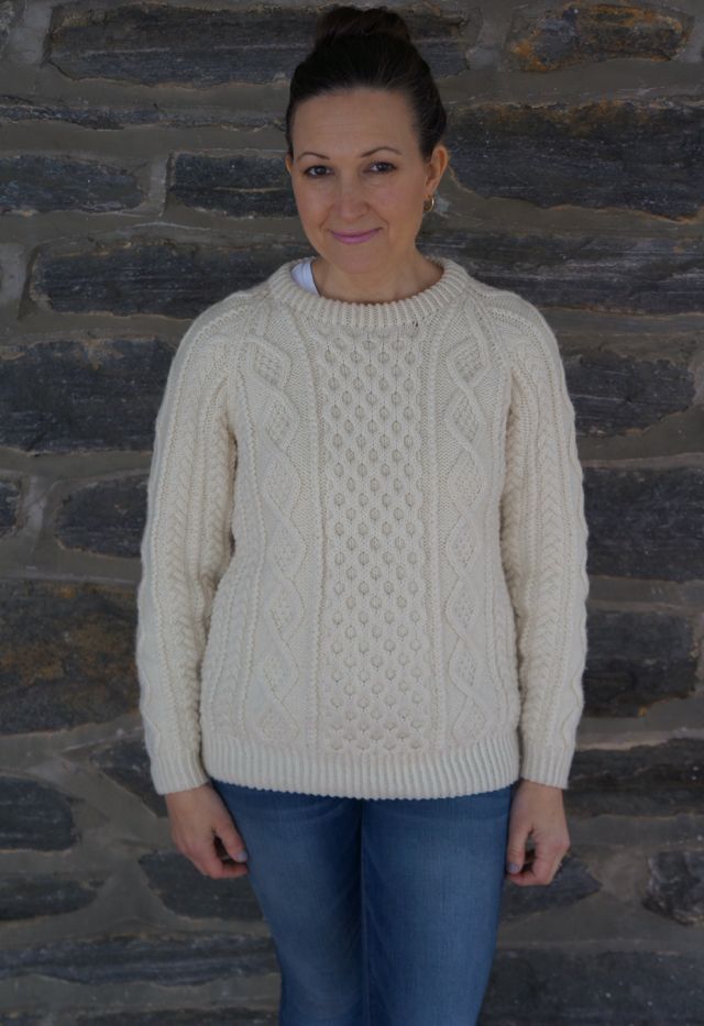 a woman standing in front of a stone wall wearing a white sweater and blue jeans