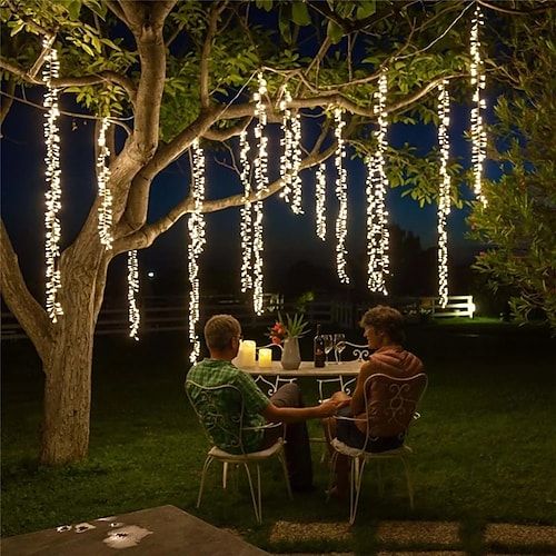 two people sitting at a table under a tree with lights hanging from it's branches