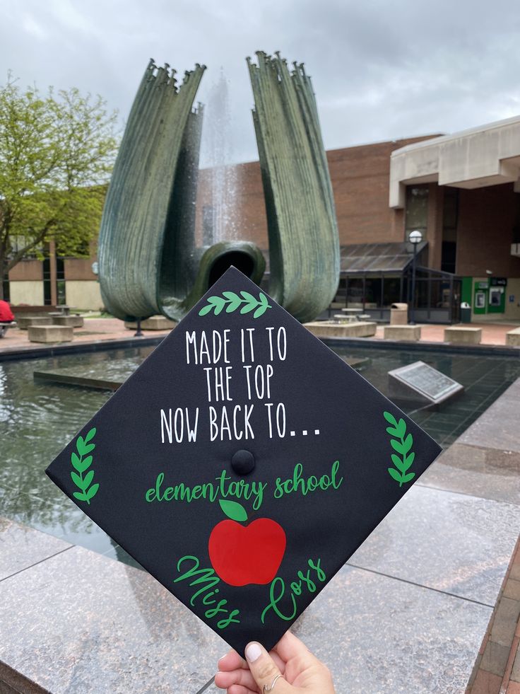 a hand holding up a black graduation cap with an apple on it and the words made it to the top now back to elementary school