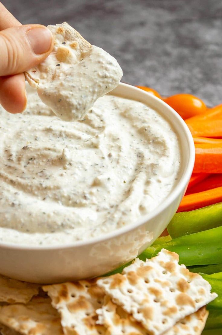 a hand dipping dip into a bowl of ranch dip surrounded by carrots and celery