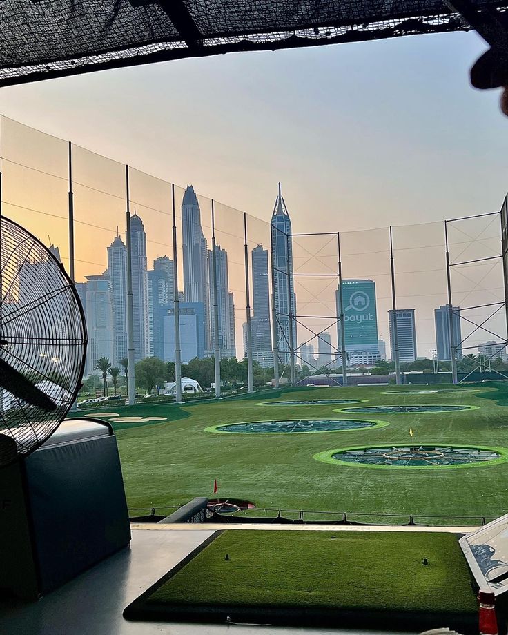 the view from inside a batting cage at a baseball field with buildings in the background