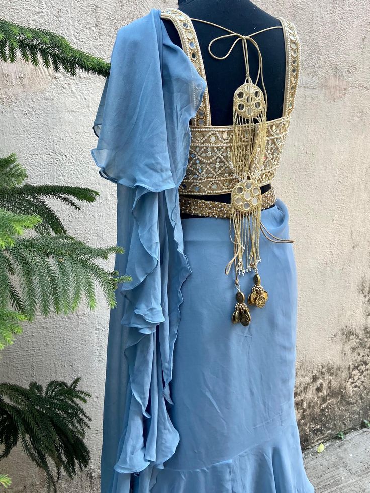 A two-piece blue ruffle saree set from the Priti Sahni collection. This beautiful blue georgette draped ruffle saree is paired with our classic mirror and pearl hand embroidered bustier blouse. The blouse has mirror-sequin tassels tie-up at the back. The outfit is completed with a heavy embroidered pearl belt.
Belt Included. Elegant Chiffon Pre-draped Saree For Festivals, Georgette Choli With Ruffles And Traditional Drape, Traditional Drape Georgette Choli With Ruffles, Chiffon Saree With Ruffles In Traditional Drape, Traditional Pre-draped Saree With Ruffles For Navratri, Ruffled Chiffon Saree In Traditional Drape, Festive Chiffon Saree With Ruffles, Party Wear Chiffon Pre-draped Saree, Elegant Pre-draped Saree With Ruffles For Navratri