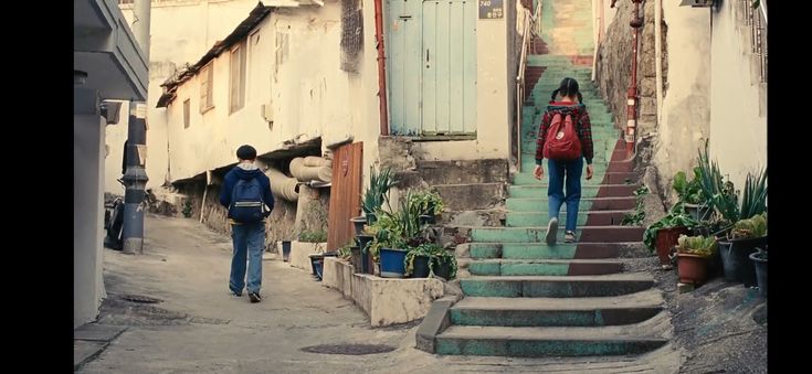 two people are walking down the street in front of some buildings with stairs and potted plants