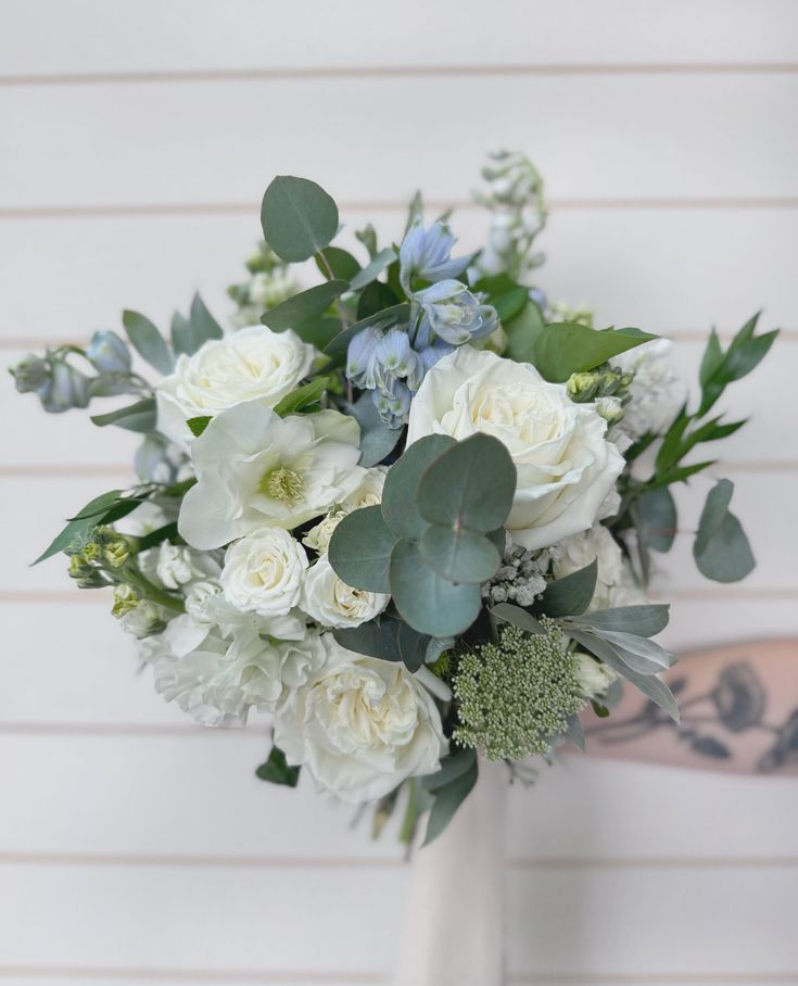 a bouquet of white flowers and greenery in front of a wooden wall with an arm tattoo