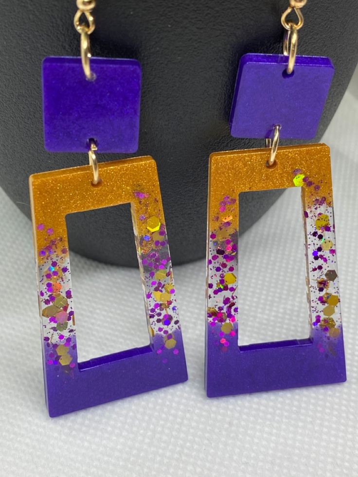 the purple and gold earrings are hanging off of a black stand on a white surface