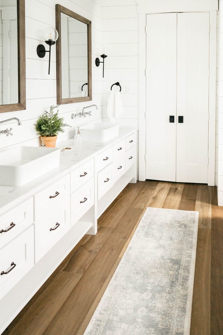 a white bathroom with two sinks, mirrors and rug on the wooden floor in front of it