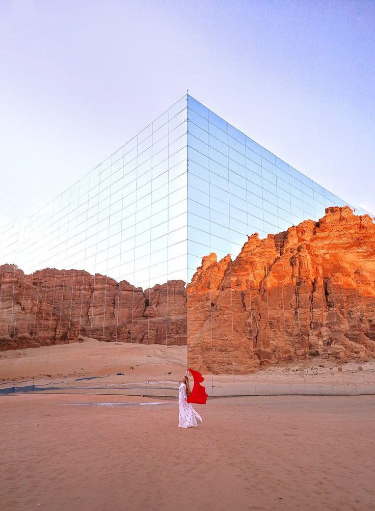 a woman walking across a sandy beach next to a tall building with a mirrored wall on it's side