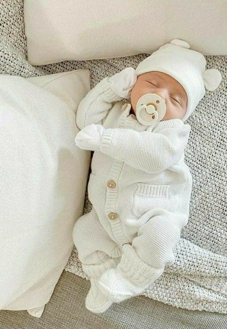 a baby in a white outfit laying on top of a bed next to a pillow