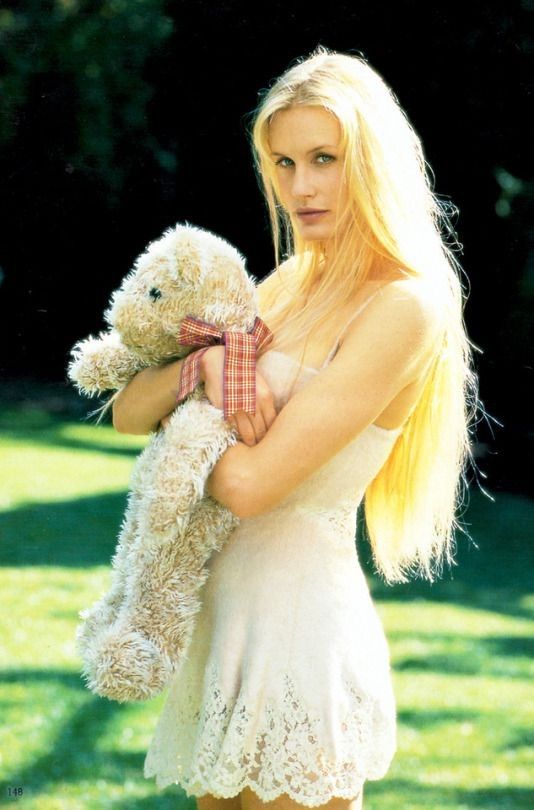 a blonde woman holding a teddy bear in her arms and posing for the camera with grass behind her