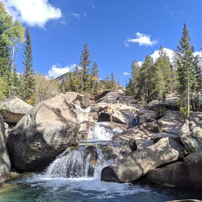 people are standing on the rocks near a small waterfall that is surrounded by pine trees