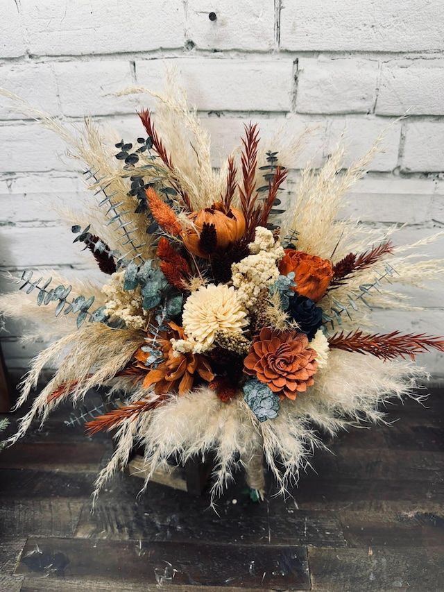 an arrangement of dried flowers and feathers in a vase on a wooden table next to a brick wall