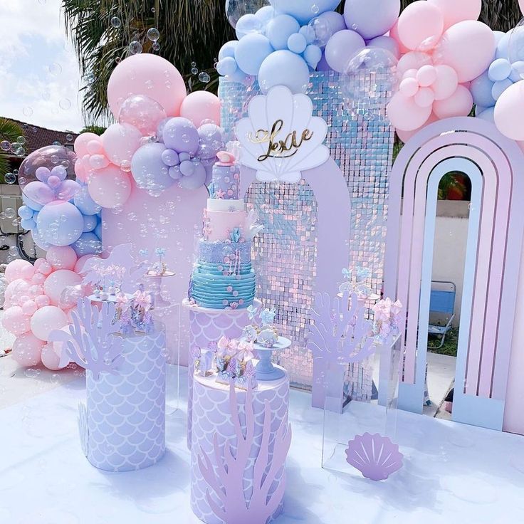 a table topped with balloons and cakes next to a wall covered in pink, blue and white