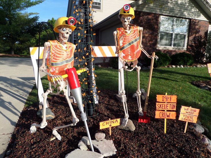 two skeleton statues in front of a house with yard signs on the ground and trees behind them