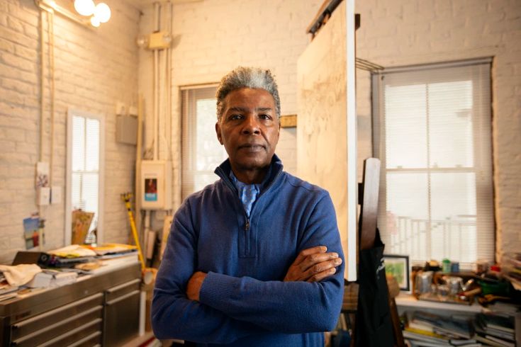 a man standing in an office with his arms crossed and looking at the camera while wearing a blue sweater