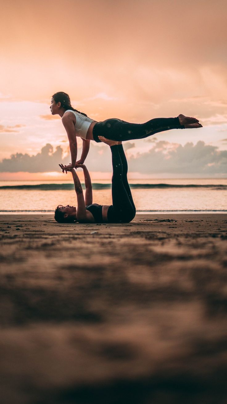 two people are doing yoga on the beach at sunset, one is holding her leg in the air