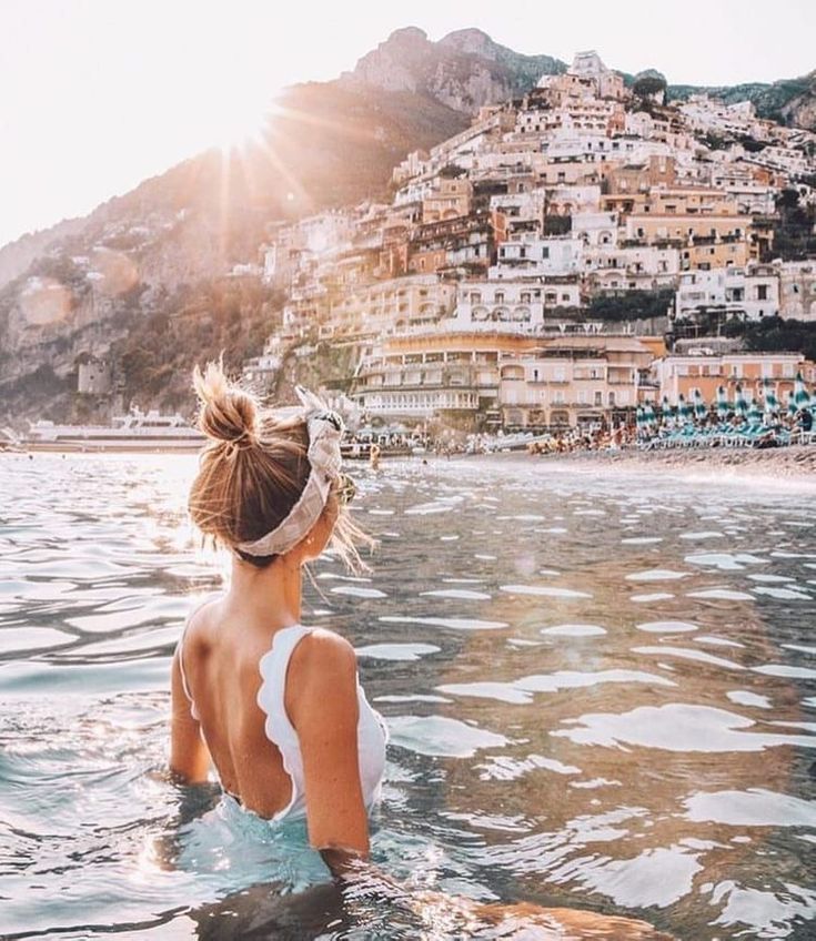 a woman is sitting in the water looking out at buildings and hills on the shore