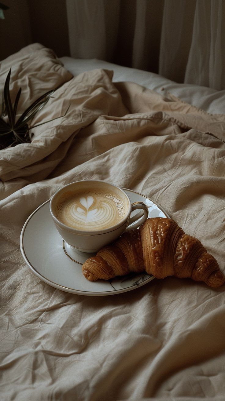 a cup of cappuccino and croissant on a bed