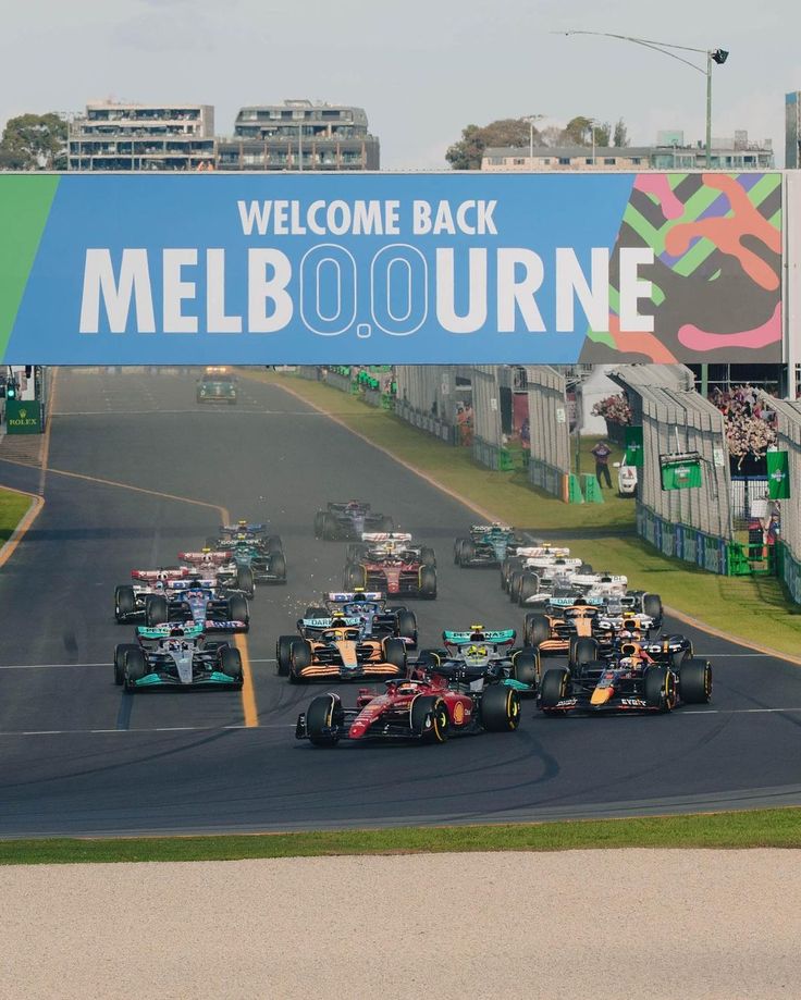 a group of cars driving down a race track with a welcome back sign in the background