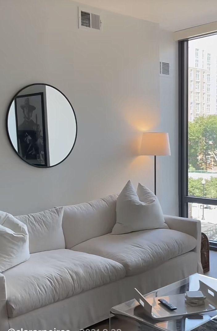 a living room filled with white furniture next to a large glass window and a mirror on the wall