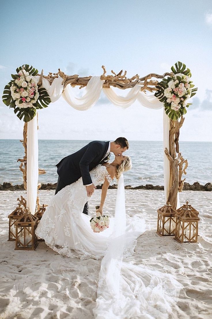 a bride and groom kissing on the beach in front of an arch decorated with flowers