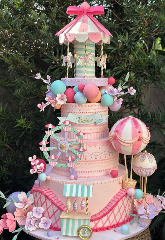 a pink and blue carousel themed cake with lots of decorations on the top, sitting in front of some trees