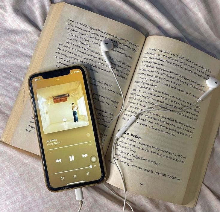 an open book and headphones are laying on a bed next to an iphone that is plugged in